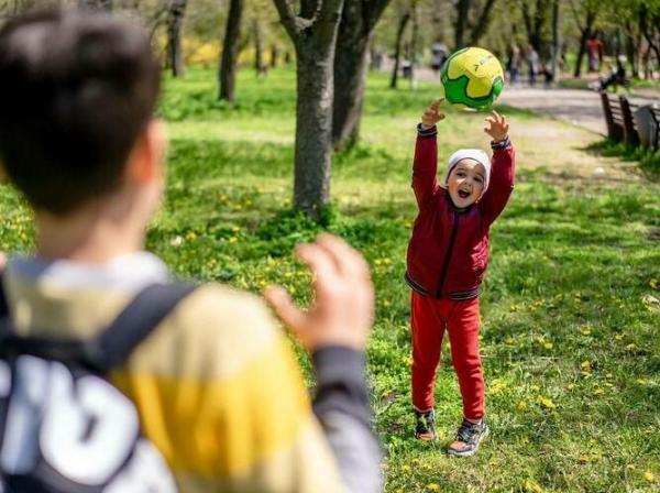 Mlajši otrok v rdečem lovi žogo, ki mu jo je vrgel fant obrnjen s hrbtom proti nam.