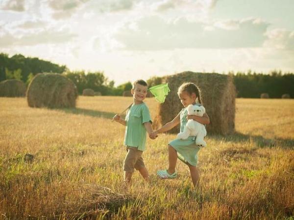 Otroka se držita za roke in stopata po travniku, zadaj bala sena. Sončen dan.