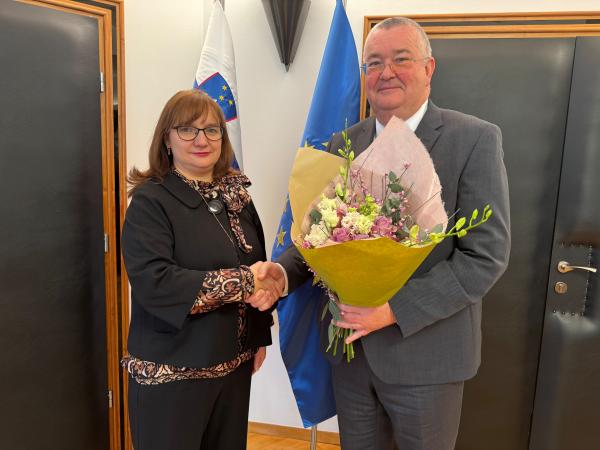 Primopredaja poslov med varuhinjo in namestnikom. Rokujeta se, namestnik v roki drži &scaron;opek rož.