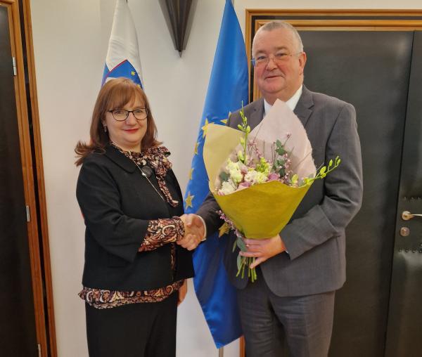 Primopredaja poslov med varuhinjo in namestnikom. Rokujeta se, namestnik v roki drži &scaron;opek rož.